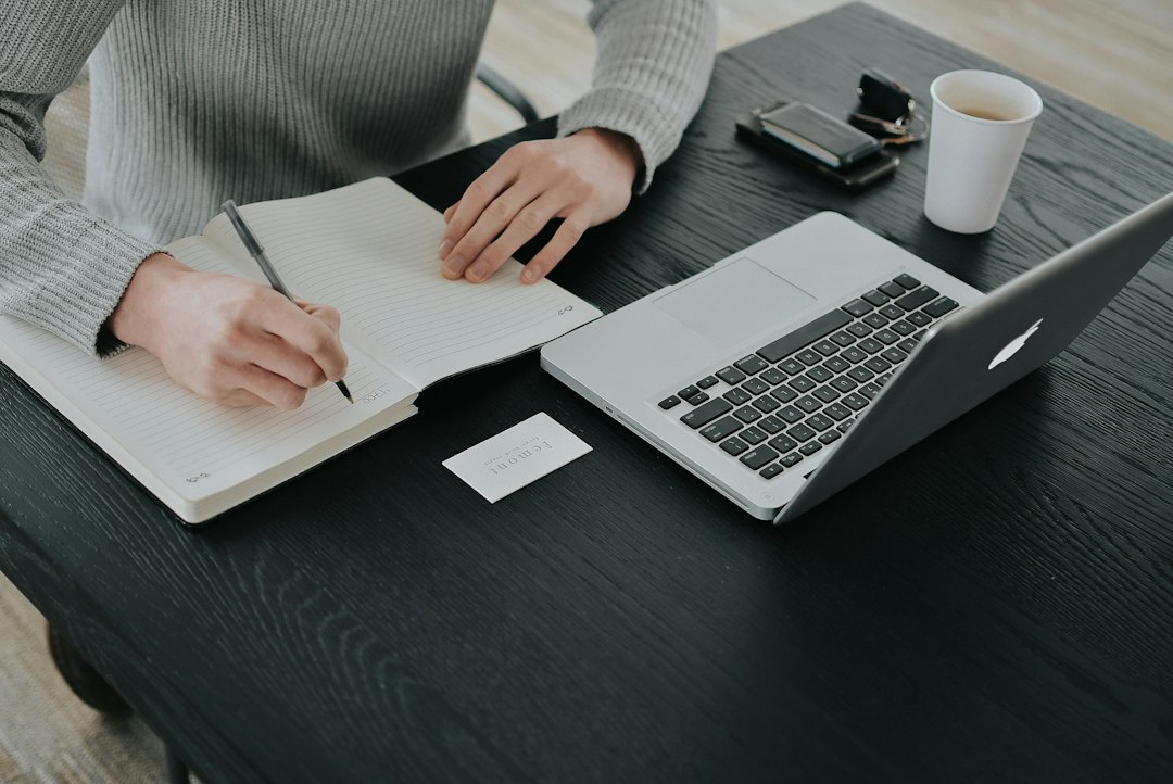 Persona che scrive su un quaderno accanto a un computer portatile su una scrivania